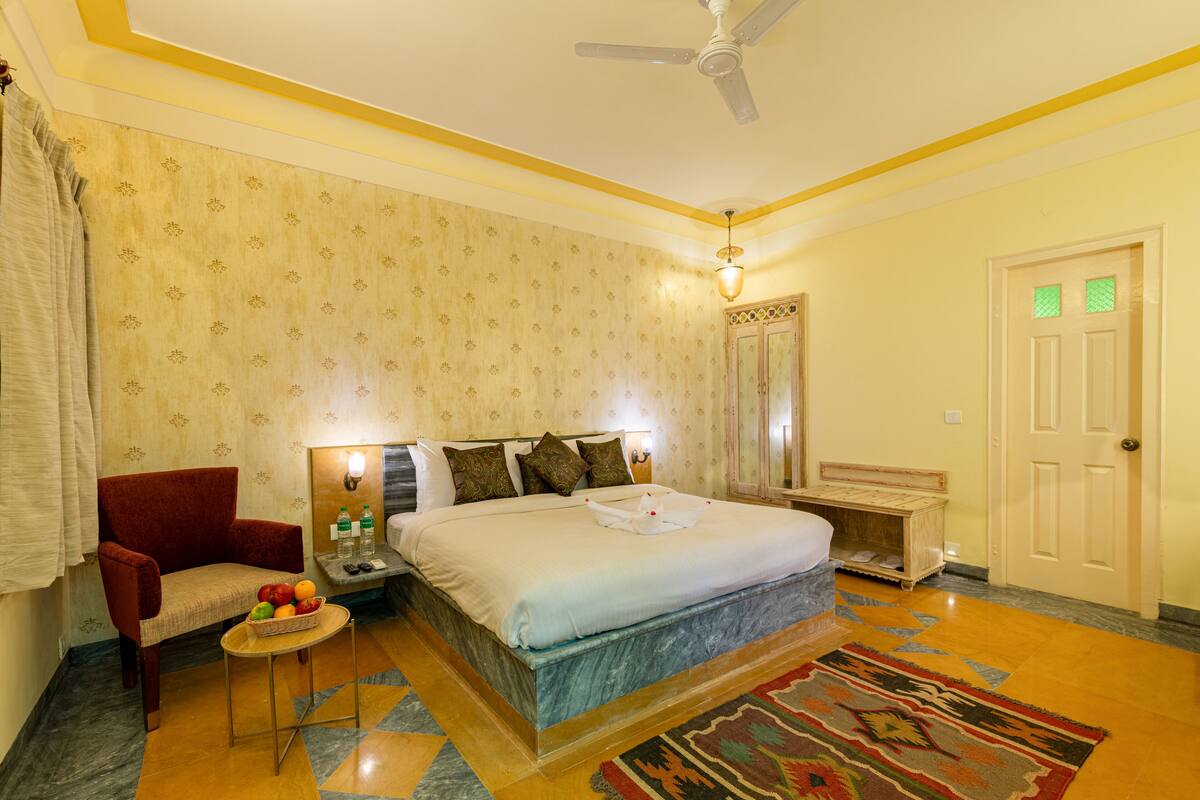 A hotel room with a bed, a chair, a small table with fruit, a nightstand, a lamp, a ceiling fan, a door, and a patterned rug.