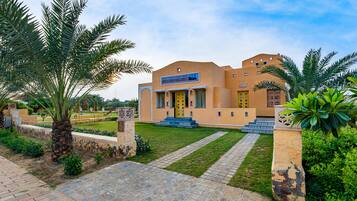 A building with a traditional architectural style, featuring a central entrance with steps, surrounded by palm trees and a well-maintained lawn.