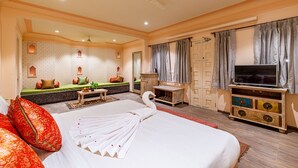 A hotel room with a large bed, a television, a seating area with a sofa and cushions, a small table with a plant, and a decorative wall with built-in lighting.