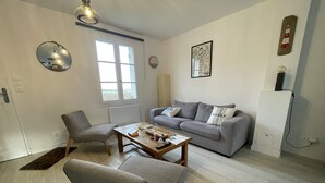 Una sala de estar moderna con un sofá gris, un sillón gris y una mesa de centro de madera. Se ve una ventana con vistas al exterior, un reloj de pared y un cuadro enmarcado.