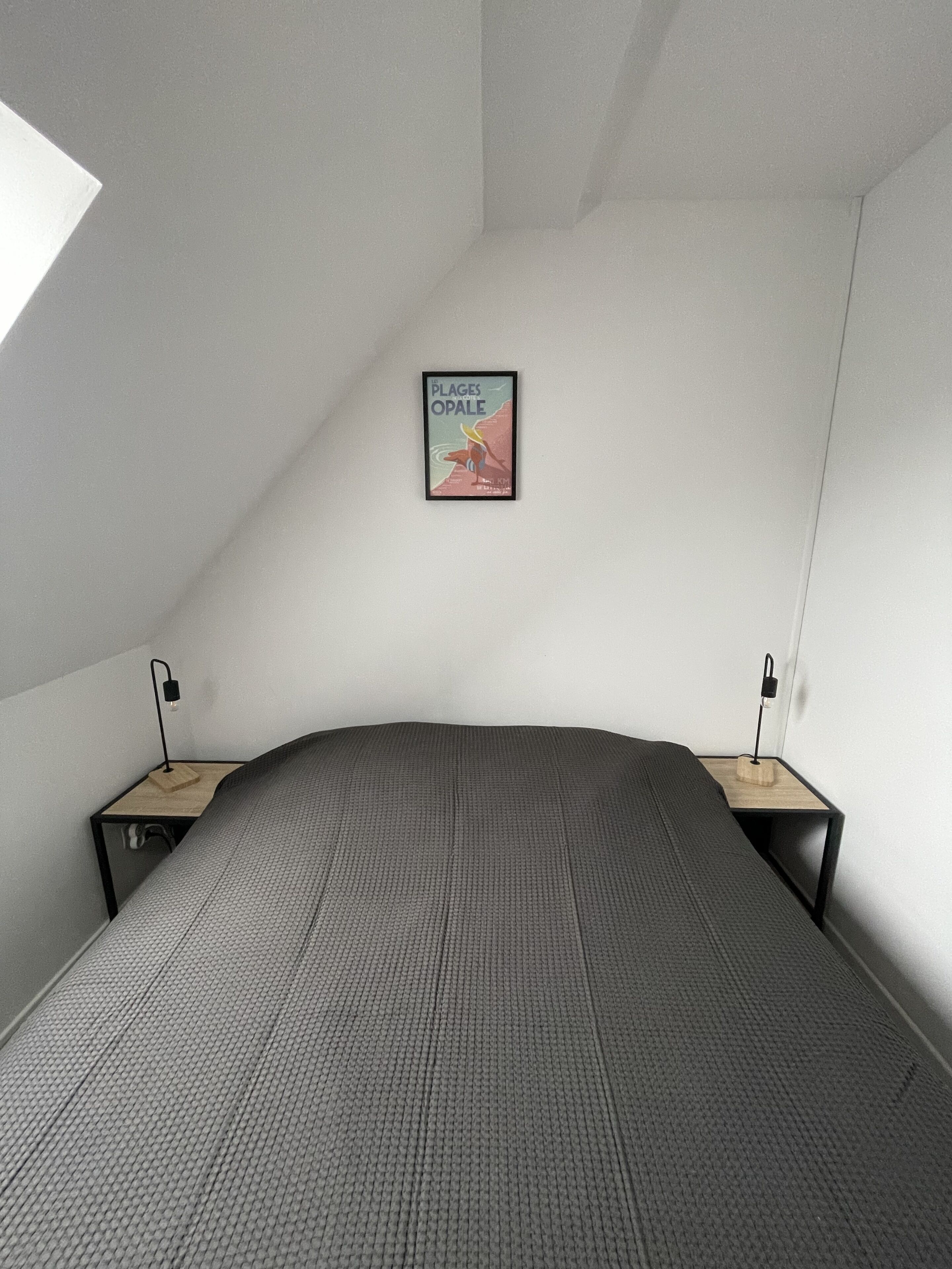 Un dormitorio con una cama grande, dos mesitas de noche con lámparas, un cartel enmarcado en la pared y un techo inclinado.