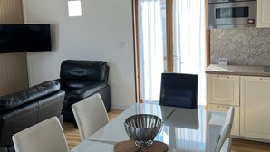 A modern living room with a leather sofa, a dining table with chairs, a TV mounted on the wall, and a kitchen area with cabinets and appliances.