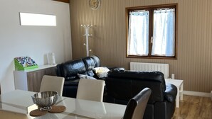 A modern living room with a black leather sofa, a dining table with chairs, a white radiator, and a window with curtains.