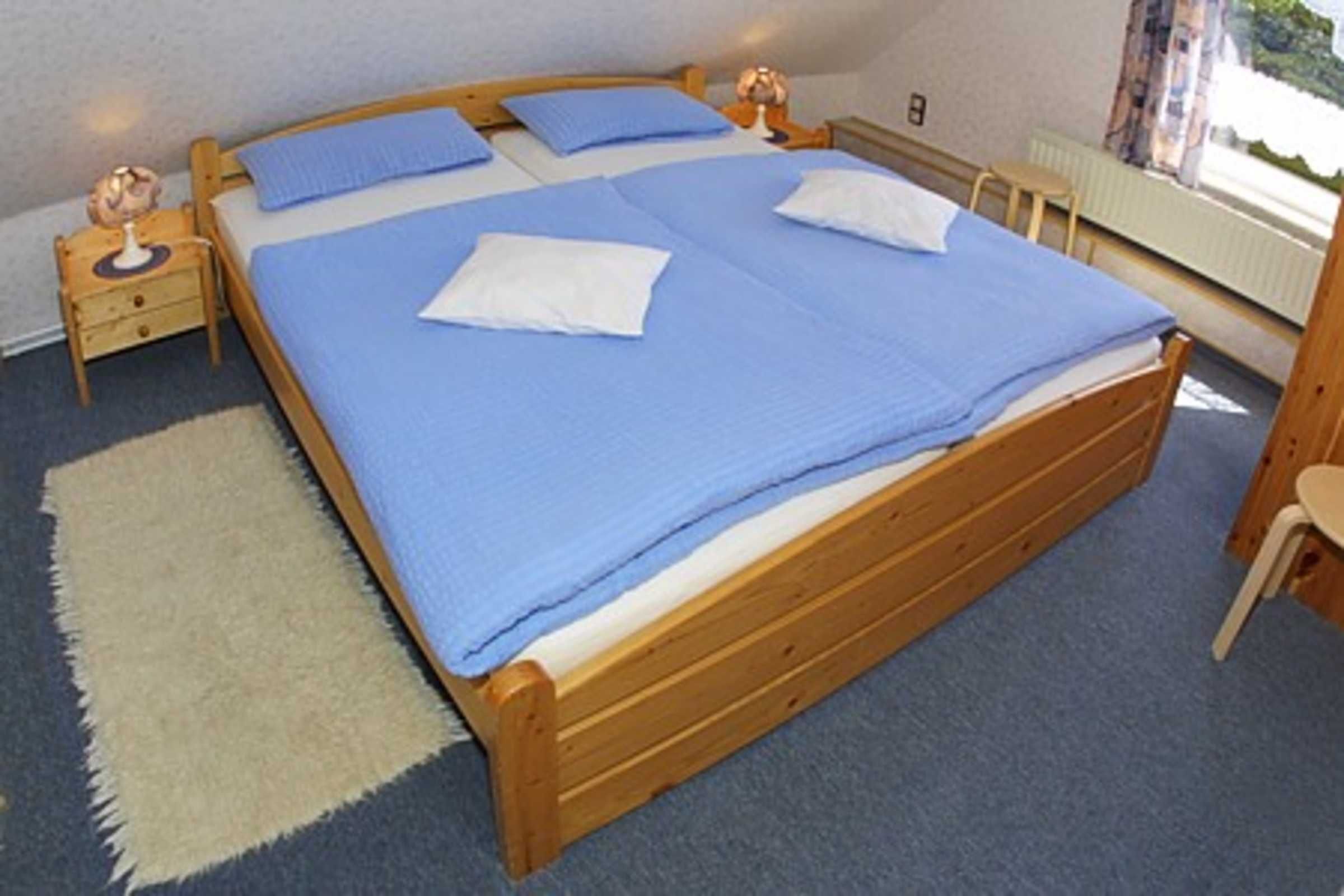 Un letto singolo con copriletto blu, due cuscini, una testiera in legno, un comodino con una lampada e una piccola finestra con tende.