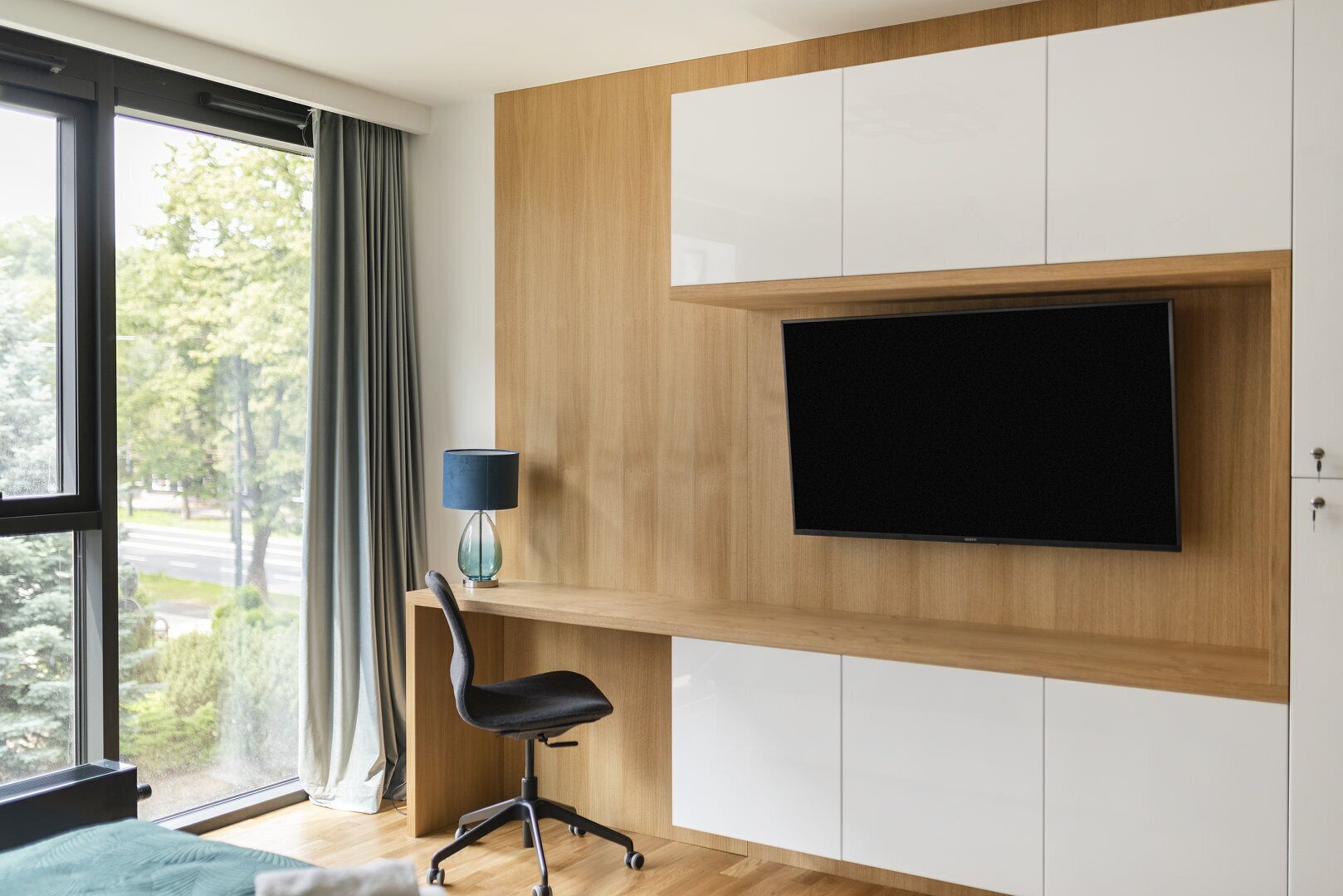 Ein modernes Zimmer mit großem Fenster, einem Flachbildfernseher an einer Holzvertäfelung, einem Schreibtisch mit Stuhl und einer Lampe.