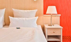 Une chambre d’hôtel avec un lit, des oreillers blancs, une table de chevet avec une lampe et un mur d’accent rouge.