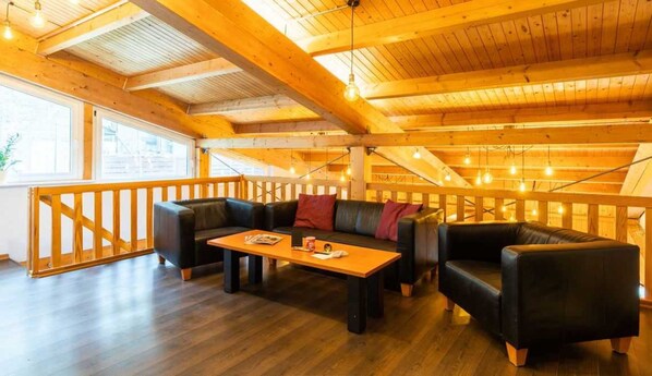 A cozy loft-style room with a wooden ceiling, a black leather sofa, a wooden coffee table, and a wooden railing.