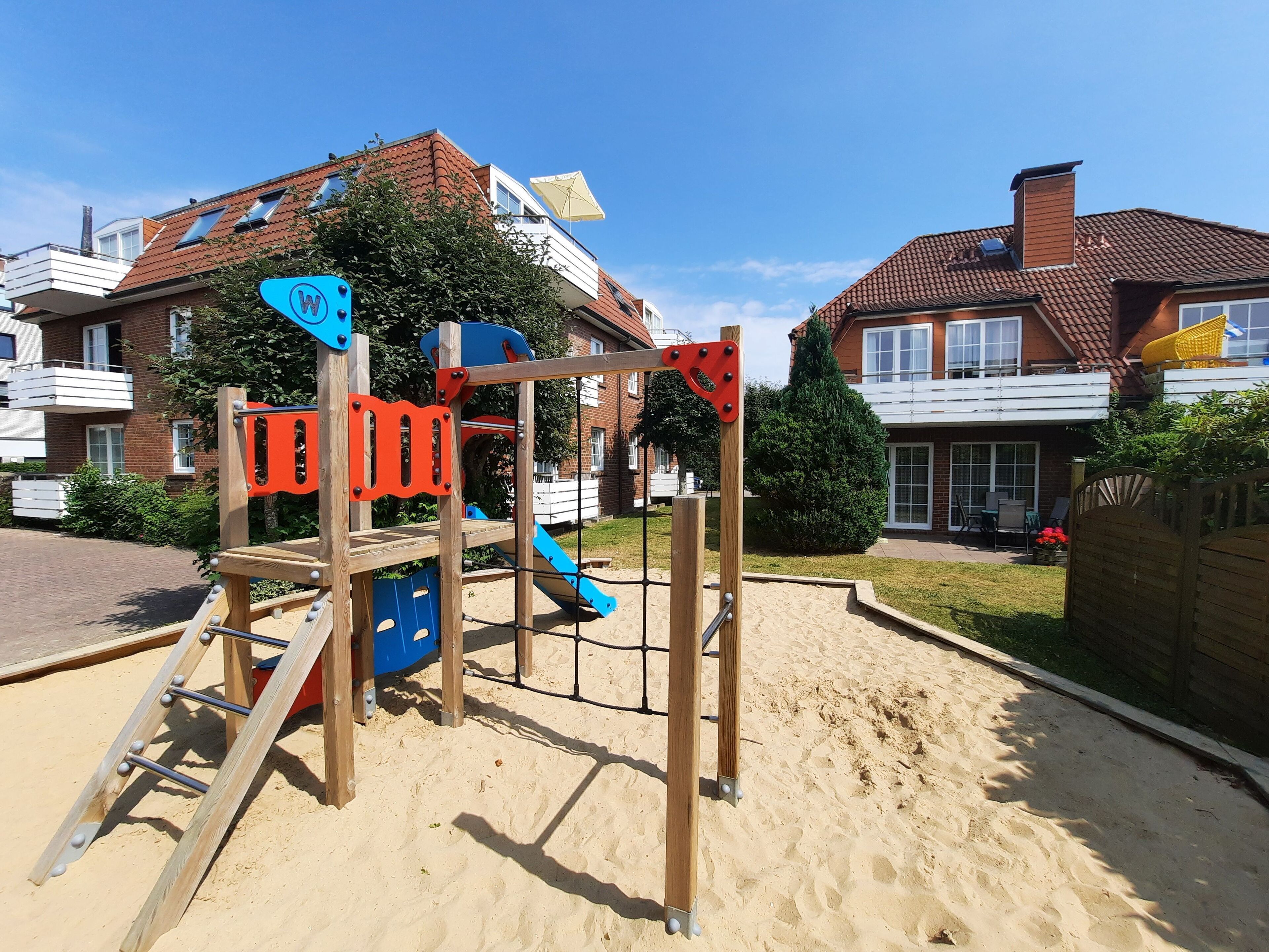 Ein Spielplatz mit einer hölzernen Kletterkonstruktion und Rutschen, umgeben von Sand, mit Häusern im Hintergrund.
