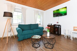 Un salon avec un canapé de couleur turquoise, un plancher en bois, une lampe sur pied, un téléviseur fixé au mur et une petite table sur laquelle est posé un vase de fleurs.