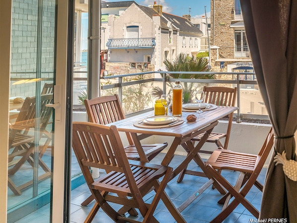 Mesa de comedor para cuatro personas, con sillas de madera, una botella de jugo de naranja y un vaso de agua en un balcón con vistas a edificios.