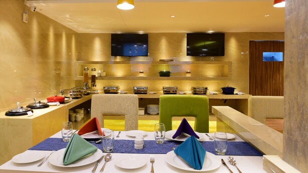 A dining area with a long table set for four, equipped with plates, cutlery, and glasses. There are two televisions mounted on the wall above the table. - Southern Crest (Chennai)