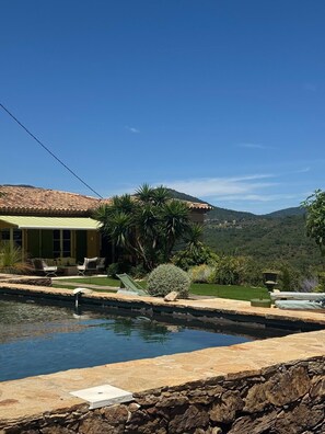 A pool with a stone border, a patio with chairs, and a house with a tiled roof.