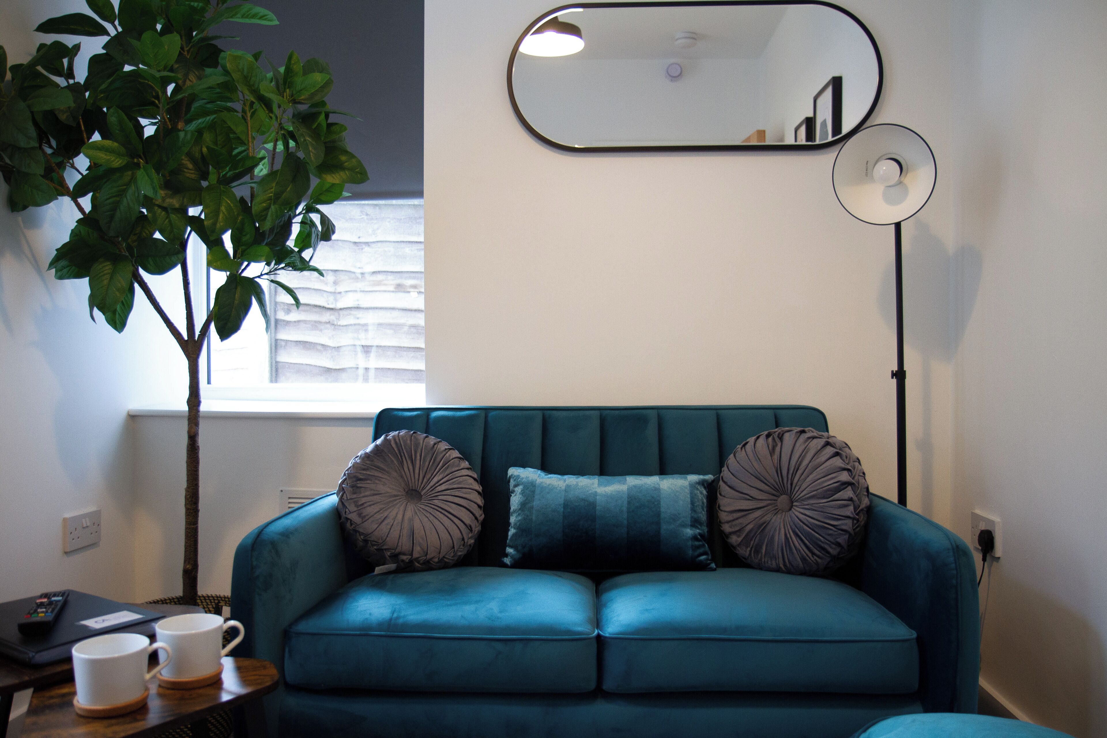 Un sofá azul verdoso con dos almohadones decorativos, una mesita con dos tazas blancas, una planta en maceta, un espejo y una lámpara de pie.