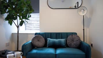 Un sofá azul verdoso con dos almohadones decorativos, una mesita con dos tazas blancas, una planta en maceta, un espejo y una lámpara de pie.