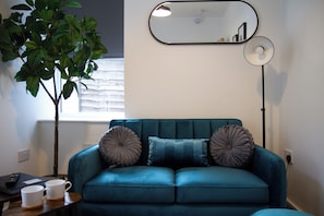 Un canapé de couleur turquoise agrémenté de deux coussins décoratifs, une petite table sur laquelle reposent deux tasses blanches, une plante en pot, un miroir et une lampe sur pied.