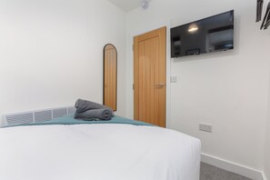 Un dormitorio con una cama, una puerta de madera, un televisor montado y una pared blanca.