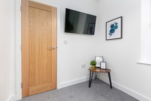 Un pasillo con una puerta de madera, un televisor de pantalla plana montado en la pared y una pequeña mesa con una planta y cuadros enmarcados.