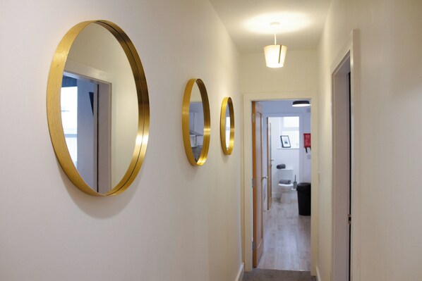 A hallway with three gold-framed mirrors on the wall, a light fixture above, and a door leading to a bathroom.