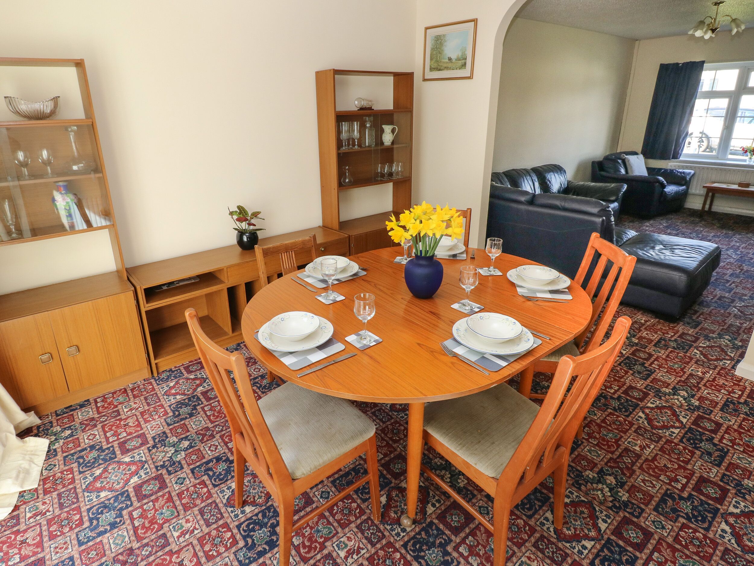 A dining area with a round wooden table set for four, surrounded by wooden chairs with grey cushions. A blue vase with yellow flowers is on the table.