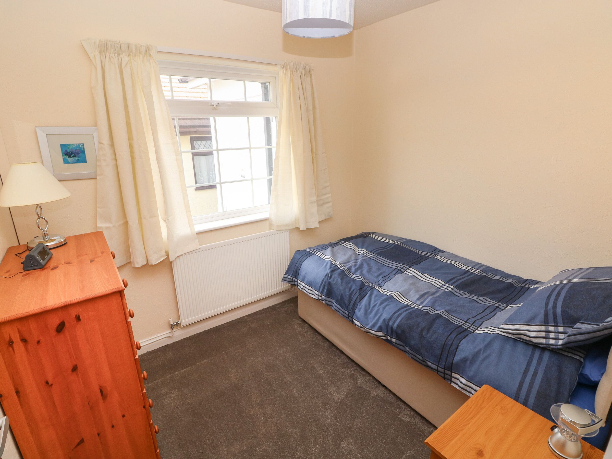 A small bedroom with a single bed, a wooden dresser, a lamp, and a window with curtains.