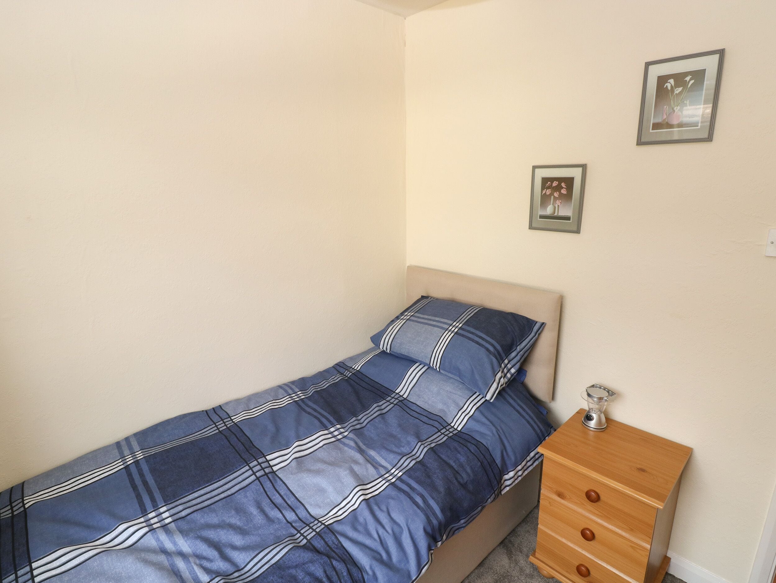 A small, single-bed room with a plain wall, a wooden nightstand with drawers, and two framed pictures.