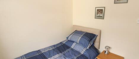 A small, single-bed room with a plain wall, a wooden nightstand with drawers, and two framed pictures.