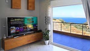 A living room with a large flat-screen TV displaying a streaming service interface, a wooden entertainment unit, and a balcony with a view of the sea. - Design Villa Nicol Heated Pool Seaview (Malevizi)