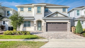 A two-story house with a garage, a front door, and a small front yard with a tree and shrubs. - Chic Home near Disney: Private Pool, Game Room & Waterpark - 320AU (Kissimmee)