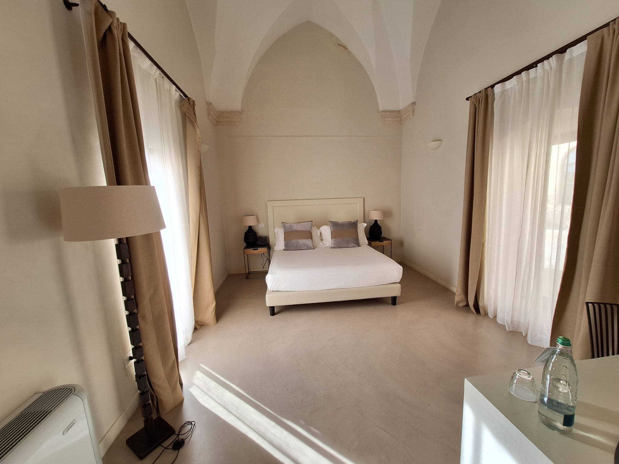 A hotel room with a large bed, two bedside tables with lamps, a radiator, and a desk with a bottle.
