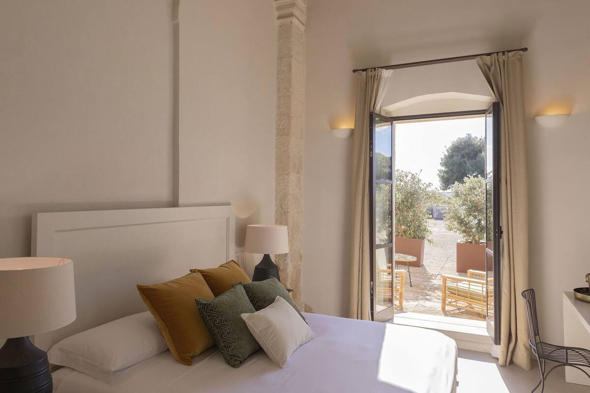 A hotel room with a bed, two bedside lamps, a view of the outdoor patio, and a window with curtains.