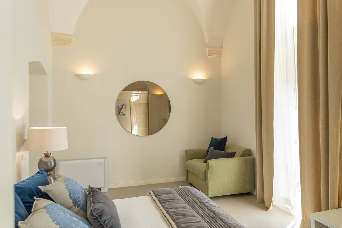 A bedroom with a bed, a green armchair, a round mirror, and a lamp.