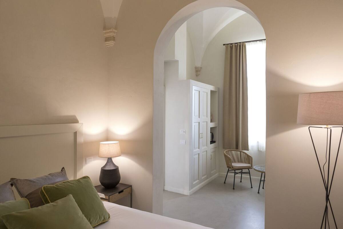 A hotel room with a bed, a nightstand with a lamp, a chair, a wardrobe, and an arched doorway leading to another room.