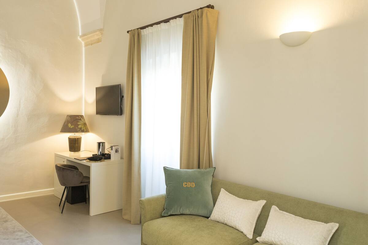 A room with a desk, a chair, a green sofa with two cushions, a lamp, and a TV mounted on the wall.