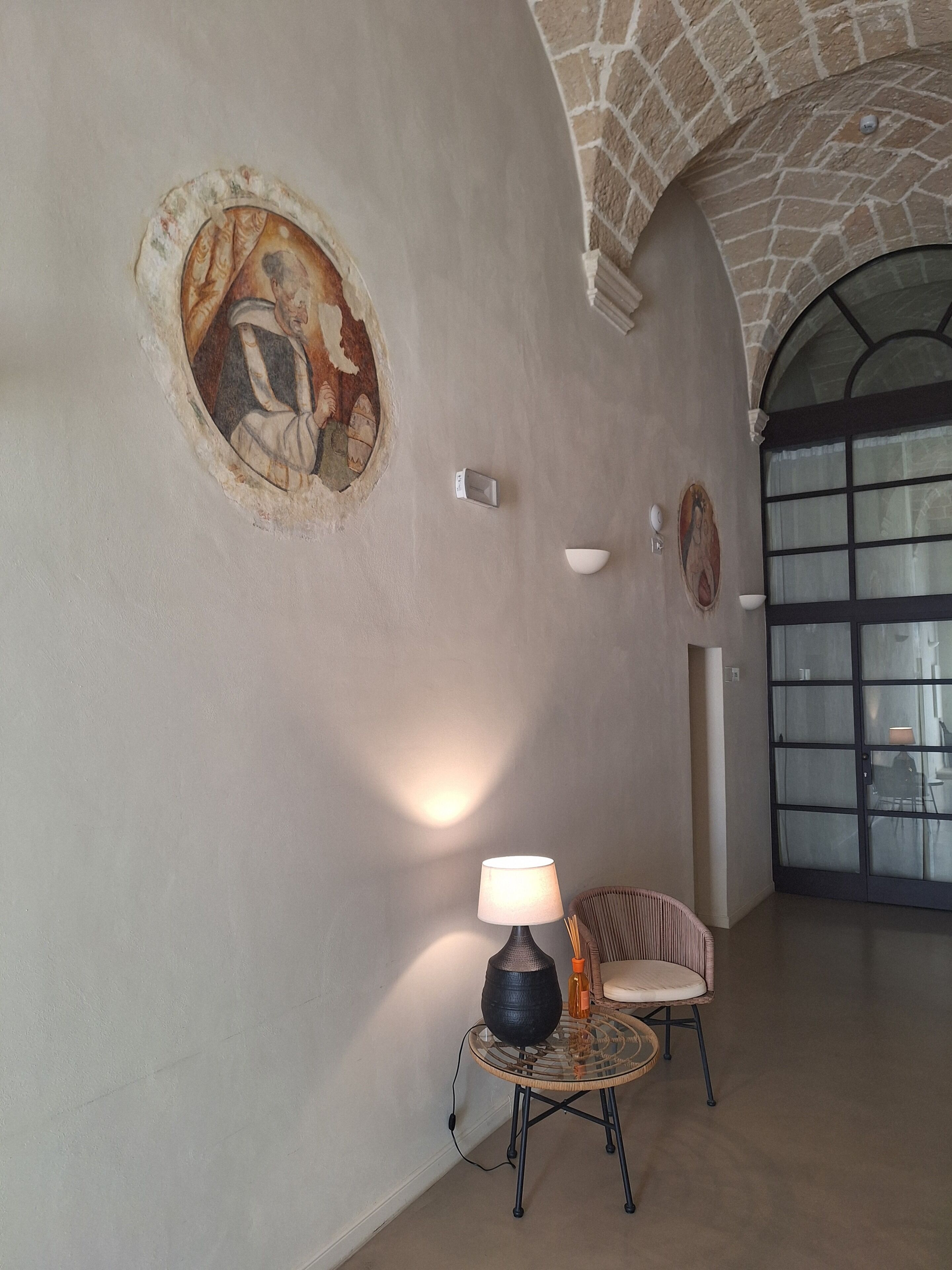 A spacious interior with a high arched ceiling, a wall with a fresco, a table with a lamp, and a chair.