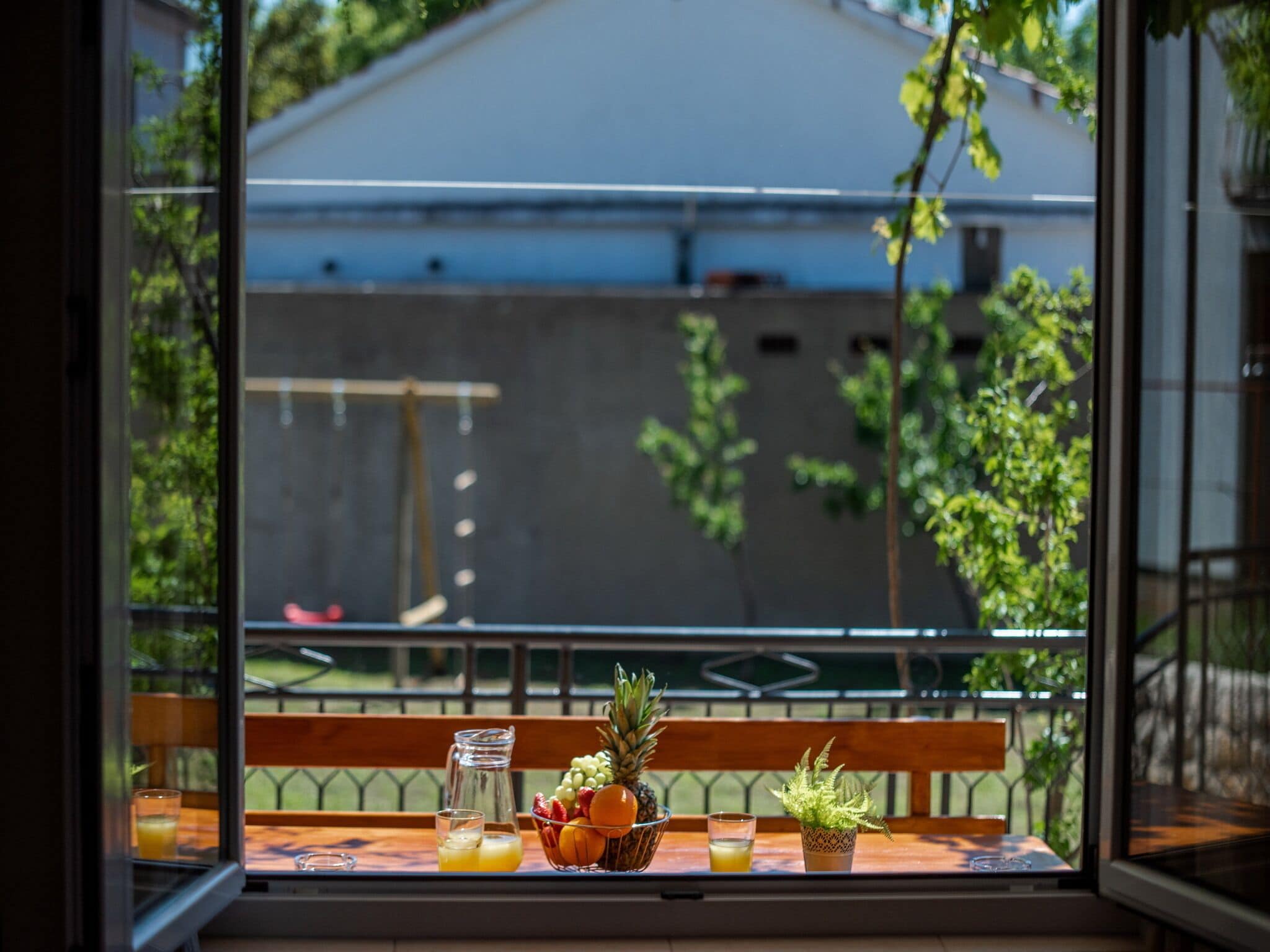 Ein Tisch mit Obstschale, Krug und Gläsern, auf einem Balkon mit Blick auf ein Gebäude und Grünflächen.