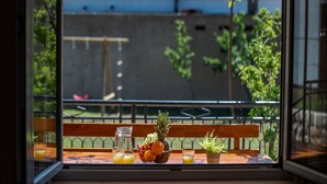 Un tavolo con una ciotola di frutta, una brocca e dei bicchieri, su un balcone con vista su un edificio e vegetazione.