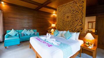 Een hotelkamer met een houten plafond, een bed met een handdoek waarop 'WELCOME' staat, een blauwgroene bank en een nachtkastje met een lamp en bloemen.