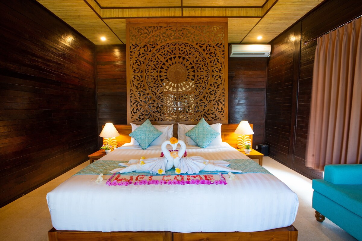 A hotel room with a large bed, wooden walls, a decorative wall panel, and a teal armchair.