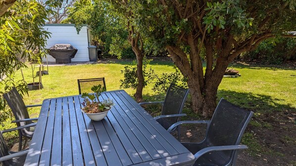 An outdoor seating area with a table and chairs, surrounded by trees and a grassy lawn.