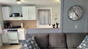 Uma cozinha compacta com armários brancos, micro-ondas e fogão. Na sala de estar, há um sofá cinza com almofadas estampadas.