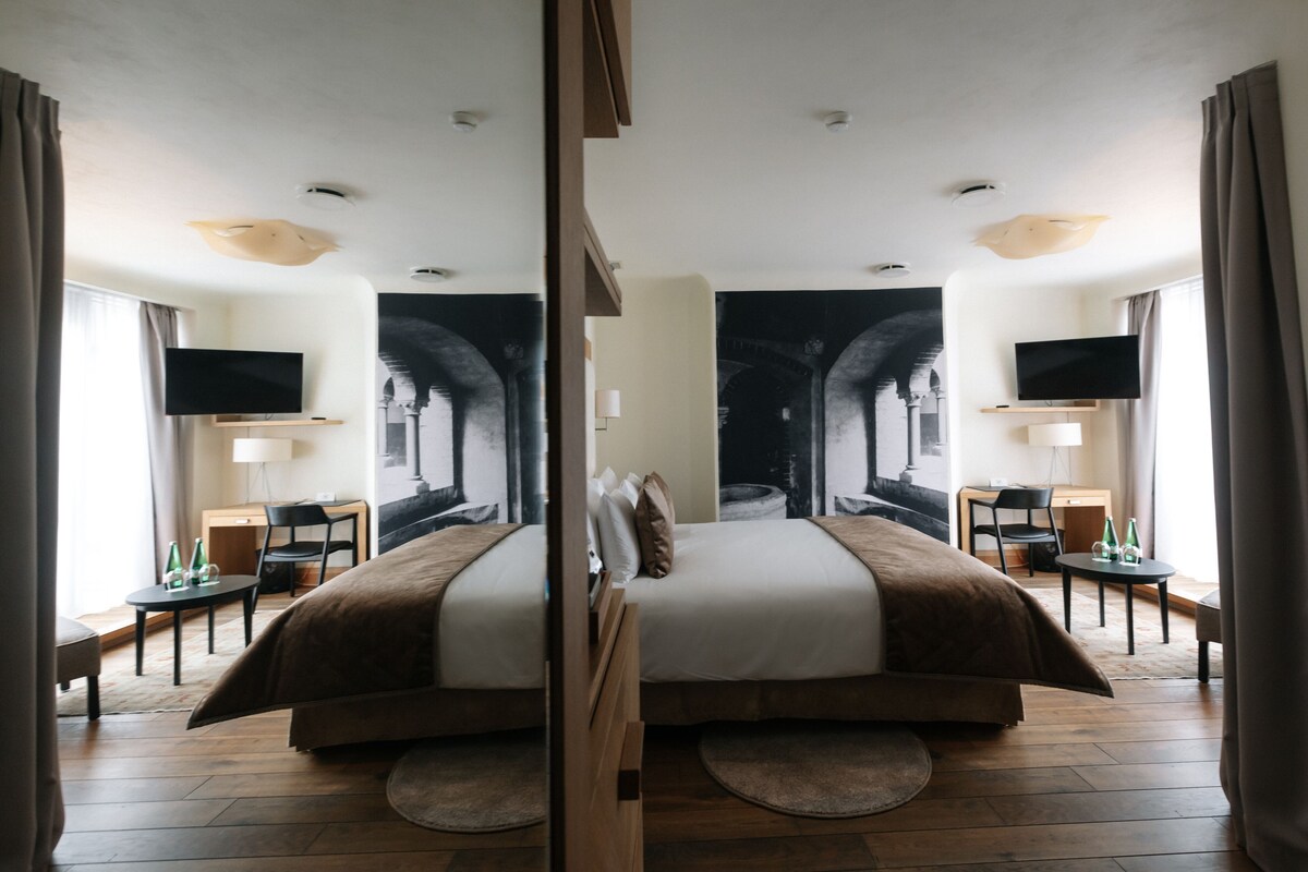 A hotel room with a large bed, two bedside tables, a flat-screen TV, and a large window with curtains.