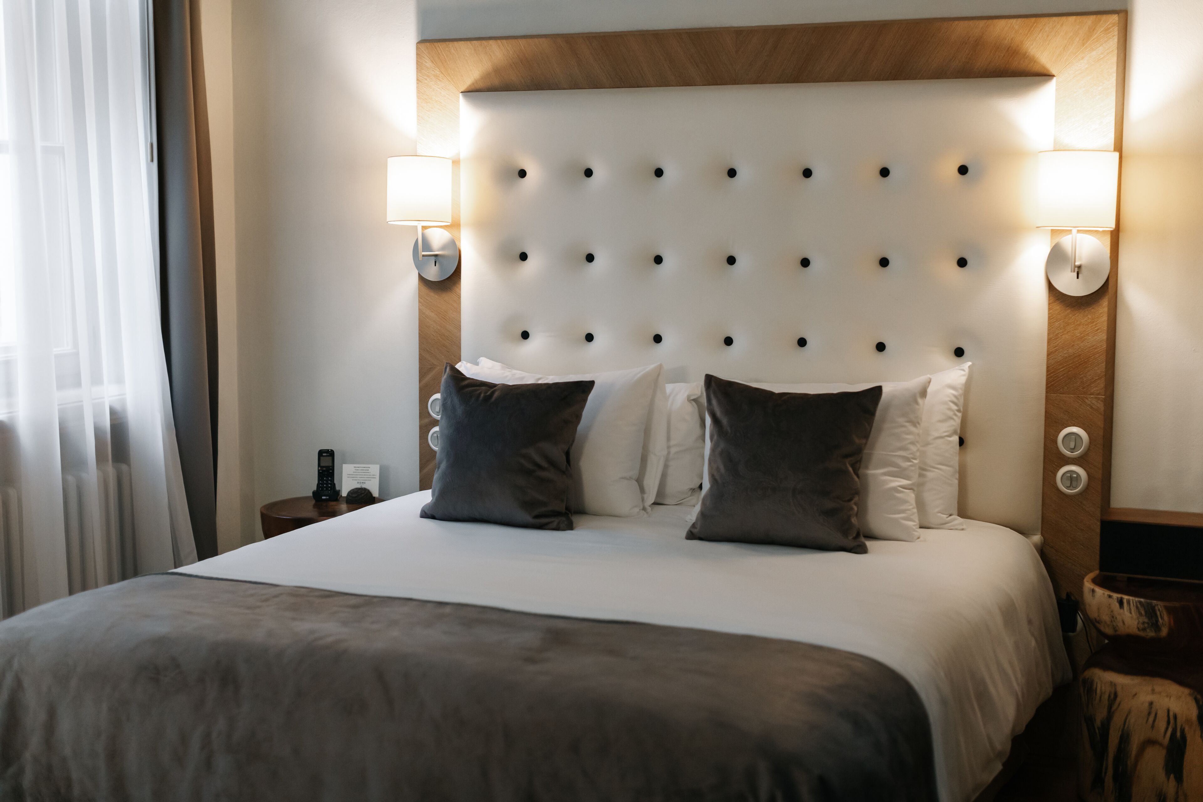 A hotel room with a tufted headboard, two bedside lamps, a bed with a grey blanket, and a nightstand with a remote control.