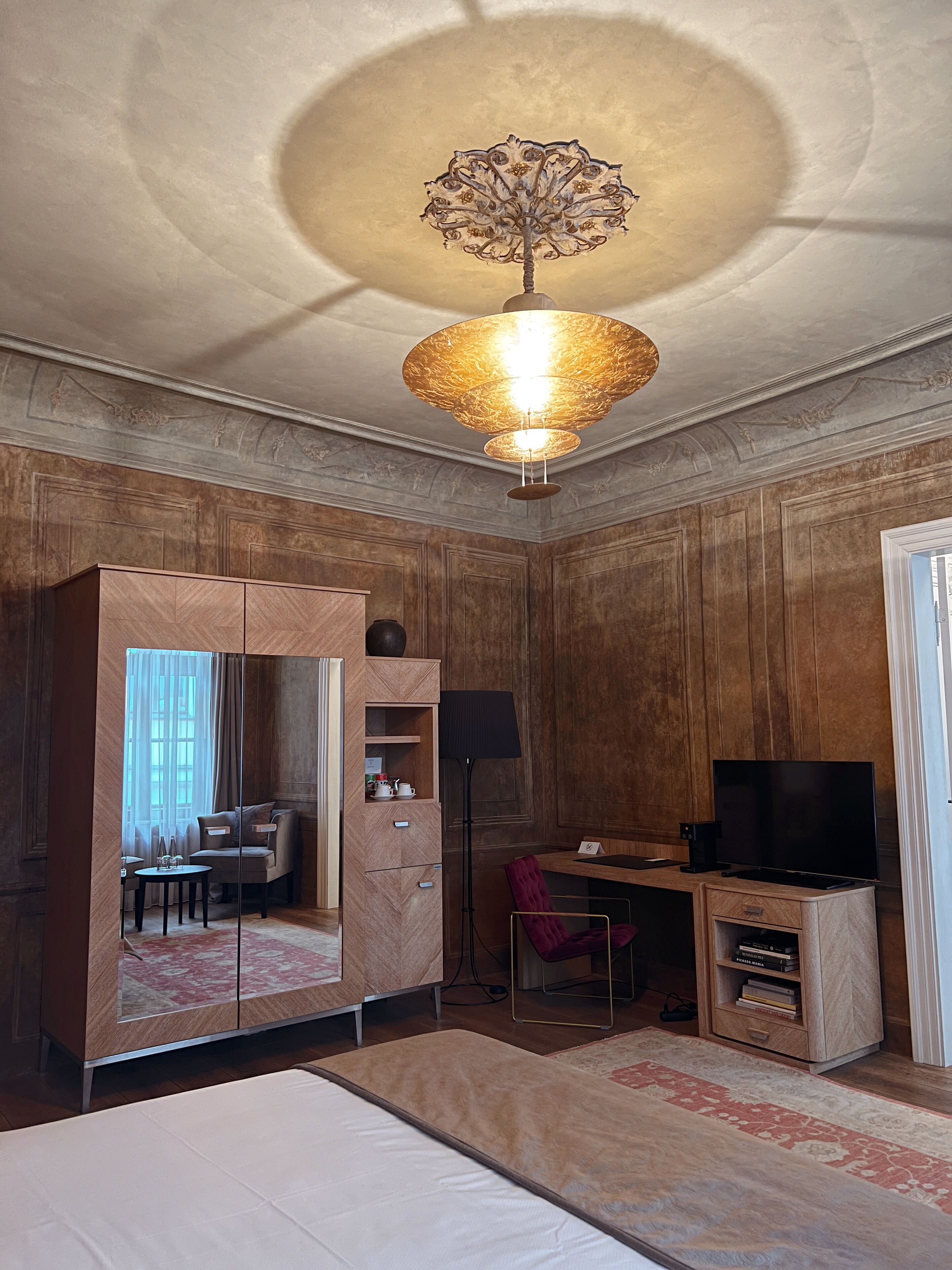 A hotel room with a large wardrobe, a desk with a flat-screen TV, a chair, a bed, and a decorative ceiling light.