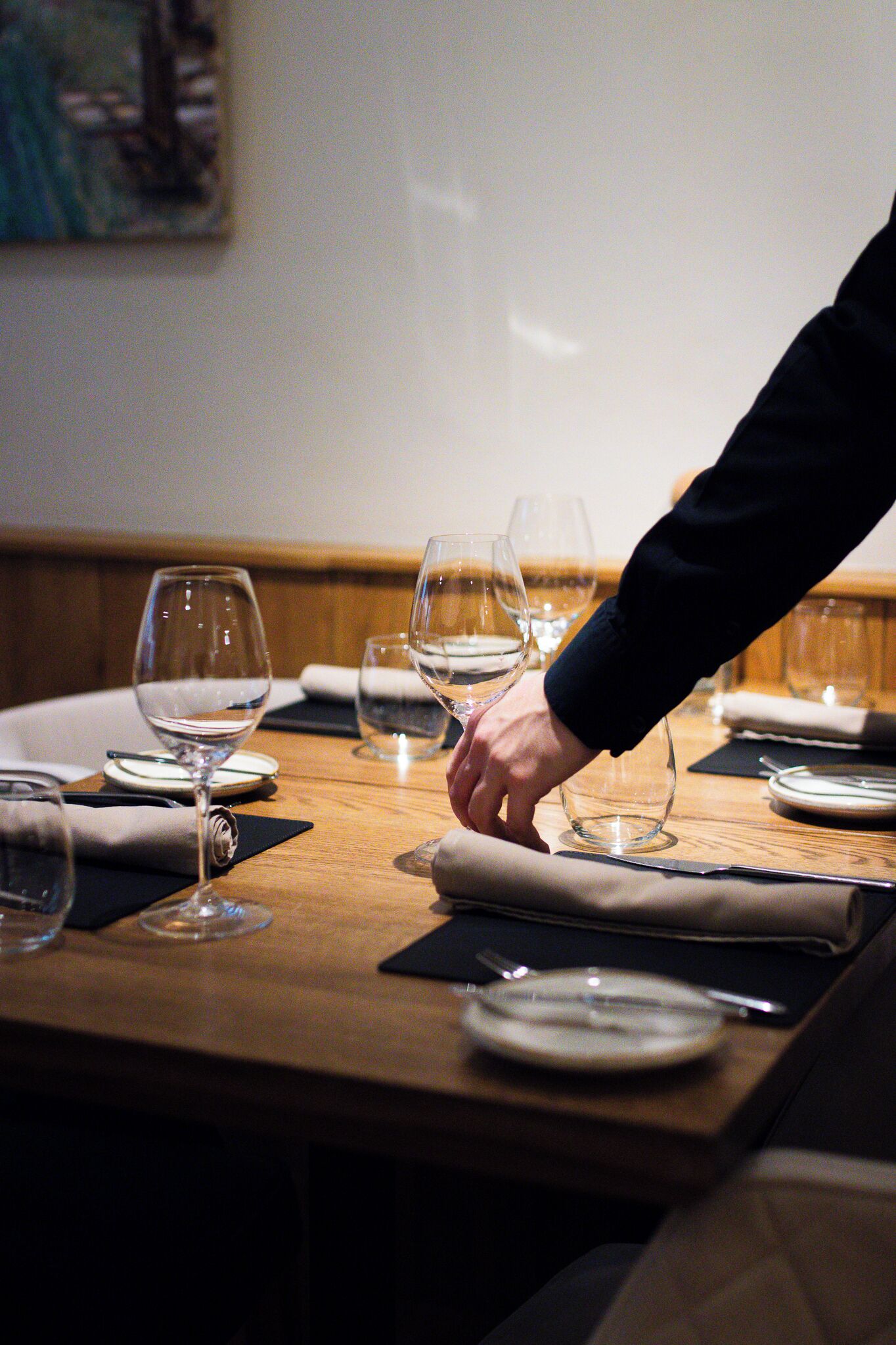 Mesa de comedor servida con copas de vino, platos y cubiertos, con la mano de alguien acomodando una servilleta.
