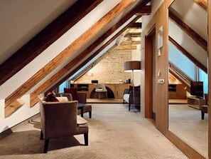 An attic room with a sloped ceiling, exposed wooden beams, a stone wall, a desk, a chair, a sofa, a lamp, and a television. - Dome Hotel (Riga)