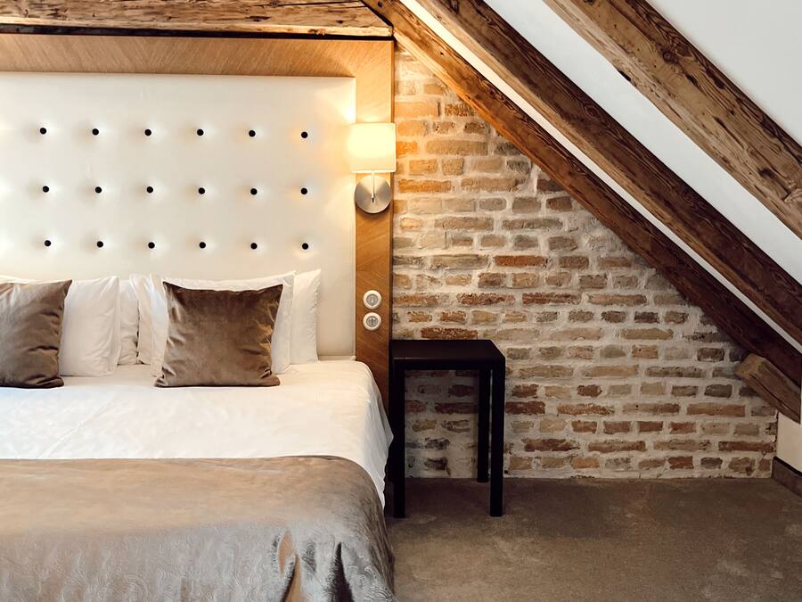 A bedroom with a tufted headboard, a bed with beige bedding, a small bedside table, and a brick wall.