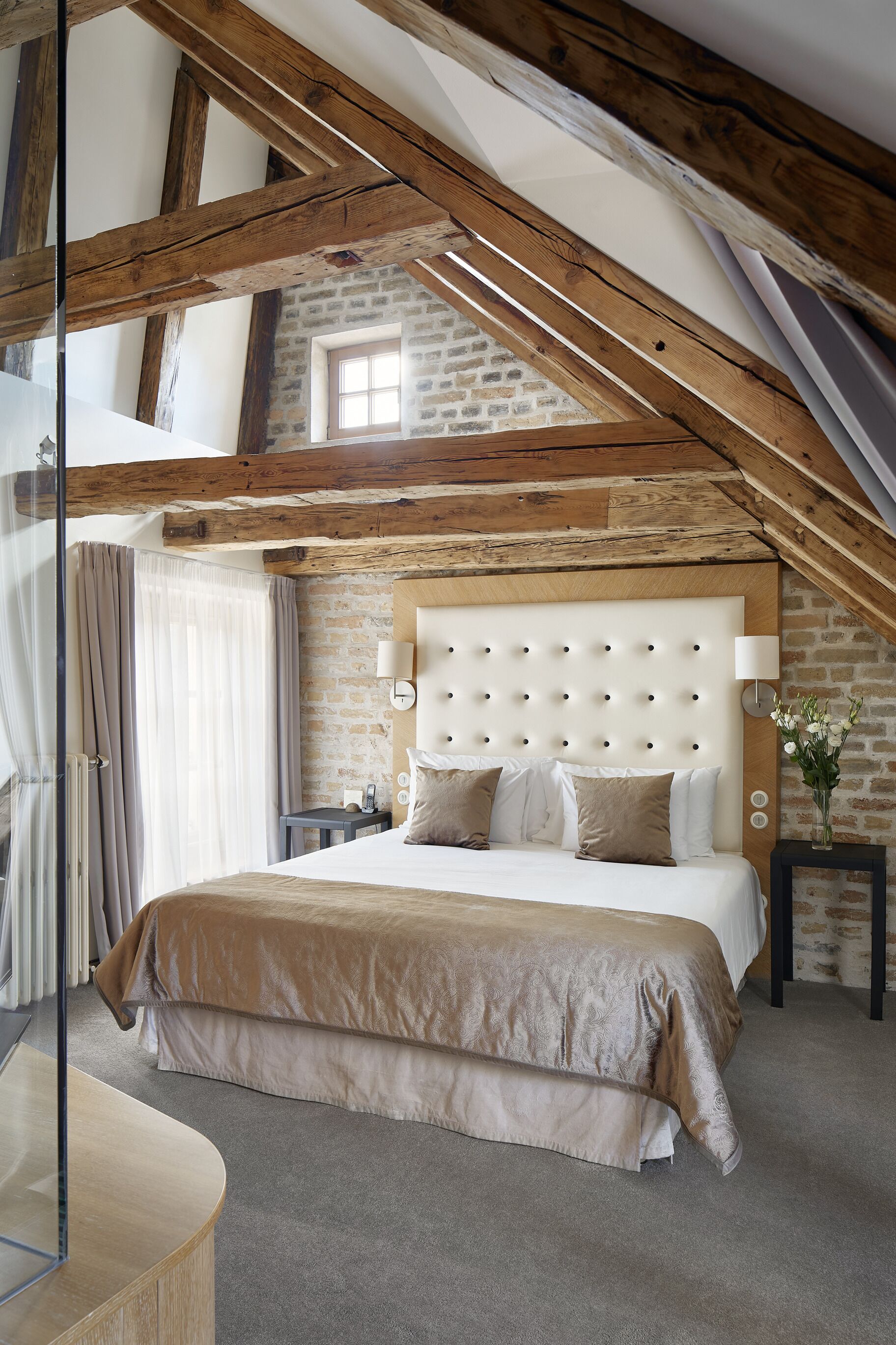 A bedroom with a high ceiling, exposed wooden beams, a brick wall, a tufted headboard, and a large window.