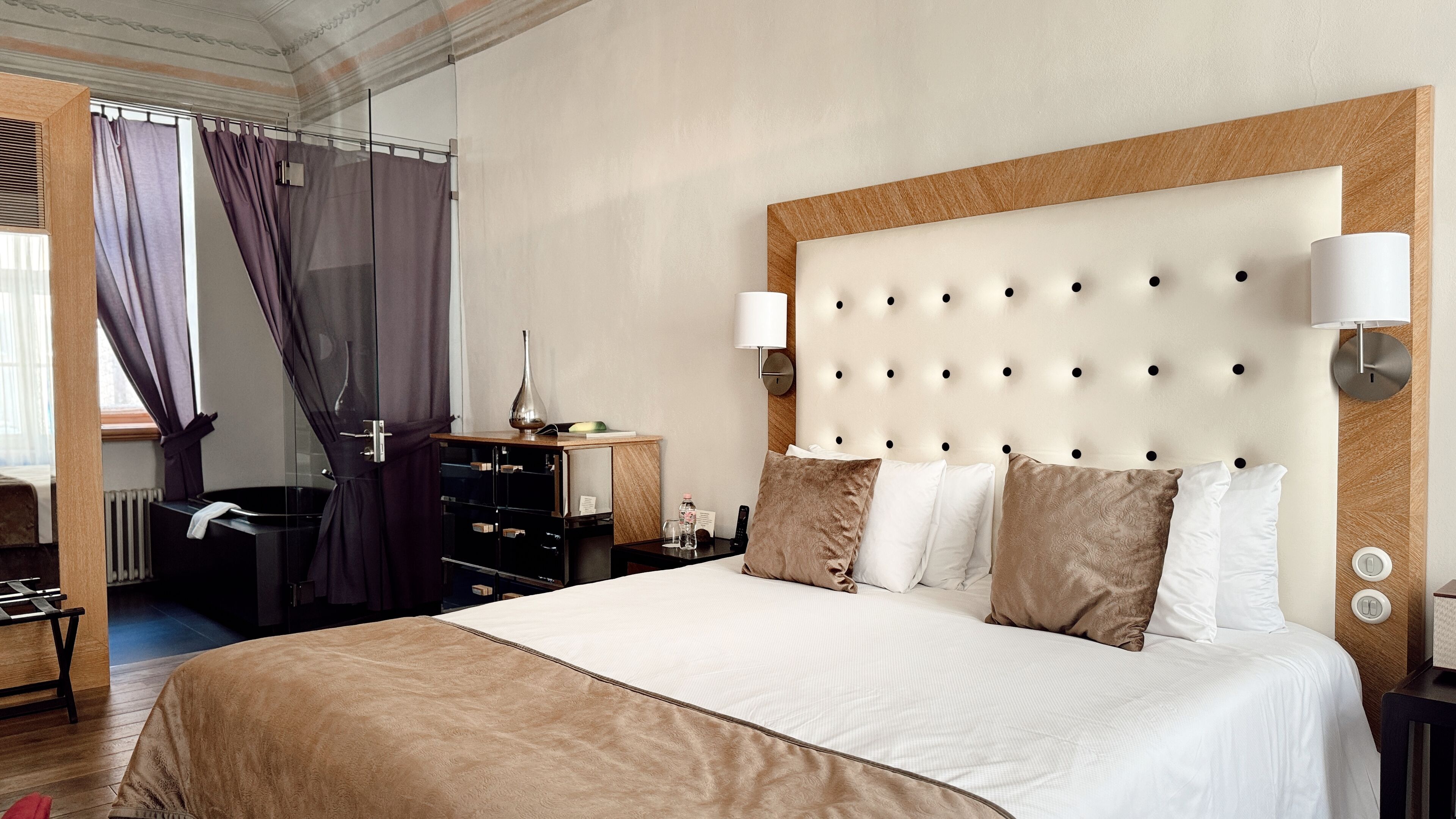 Une chambre d’hôtel avec un grand lit, une tête de lit en bois, une table de chevet avec une lampe, une commode et une fenêtre avec des rideaux.