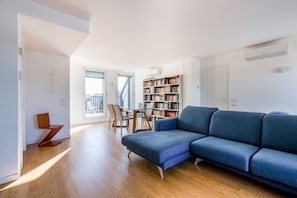 Un salon moderne avec un canapé bleu, un plancher en bois, une bibliothèque et un coin repas comprenant une table et des chaises.