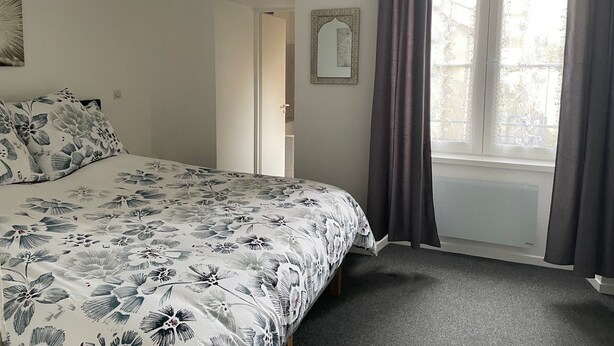 A bedroom with a bed, a window with curtains, a door, and a wall-mounted mirror.